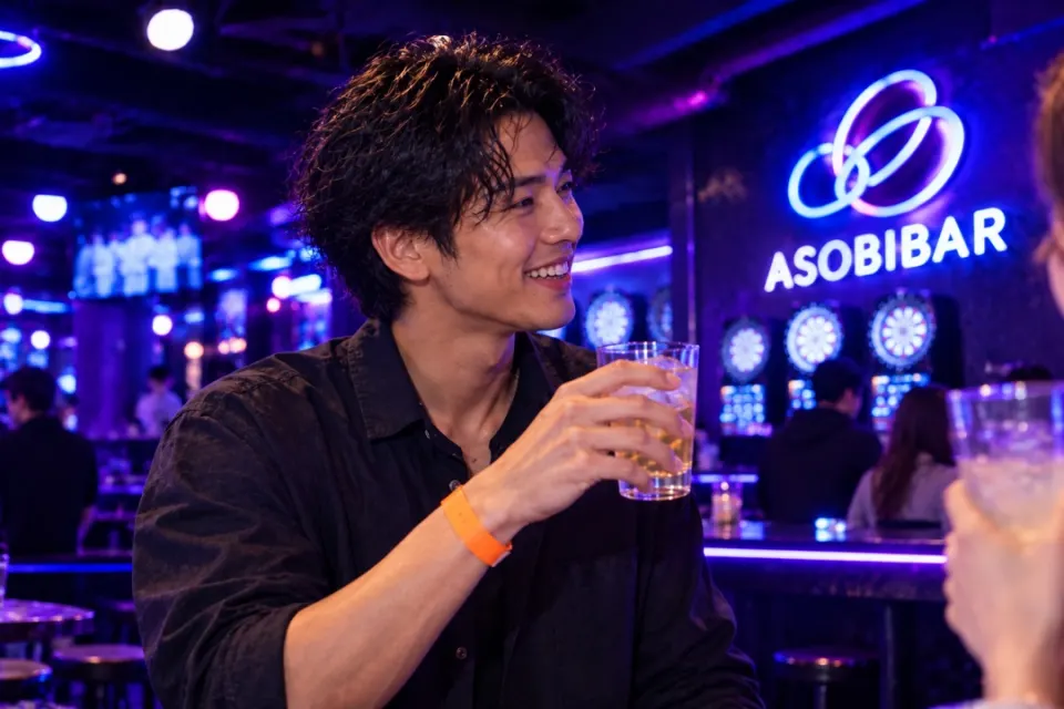 手首にオレンジのバンドを着けた男性が、ASOBIBARの店内でグラスを掲げて笑っている様子